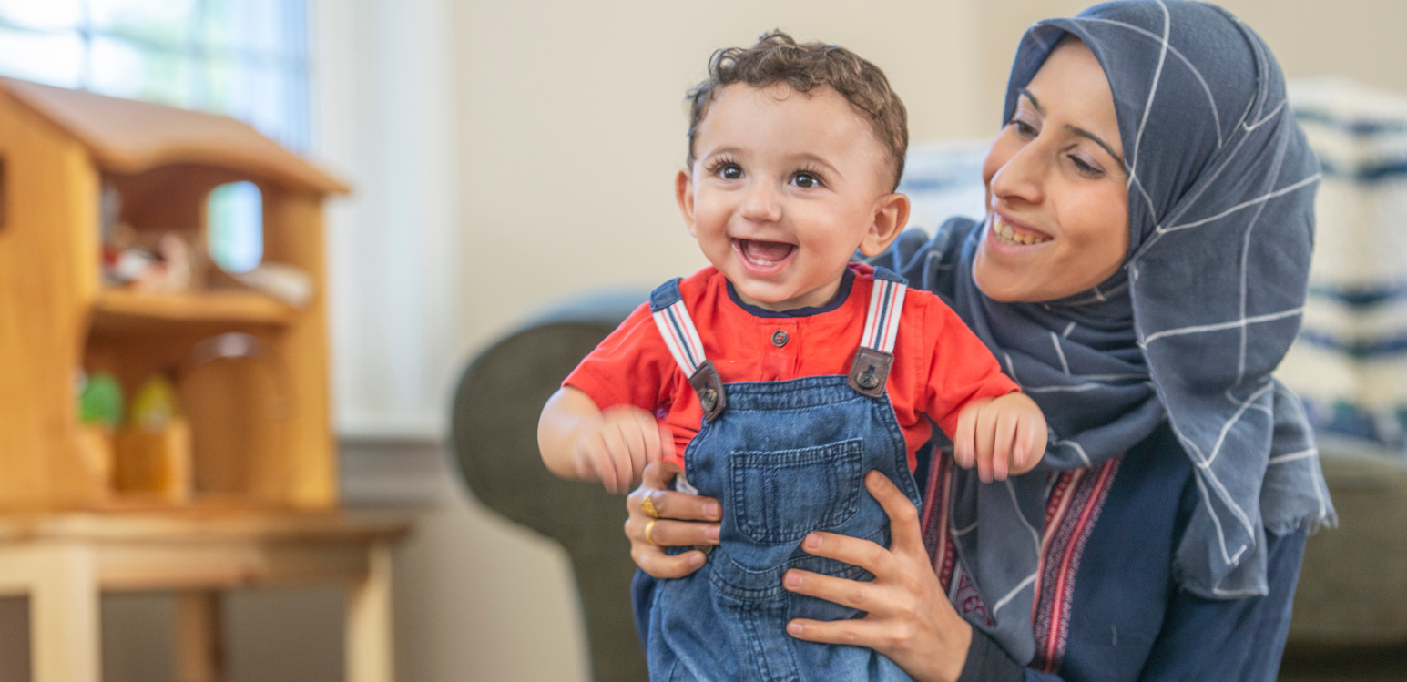 A woman wearing a hijab smiles and hugs a young boy wearing overalls.