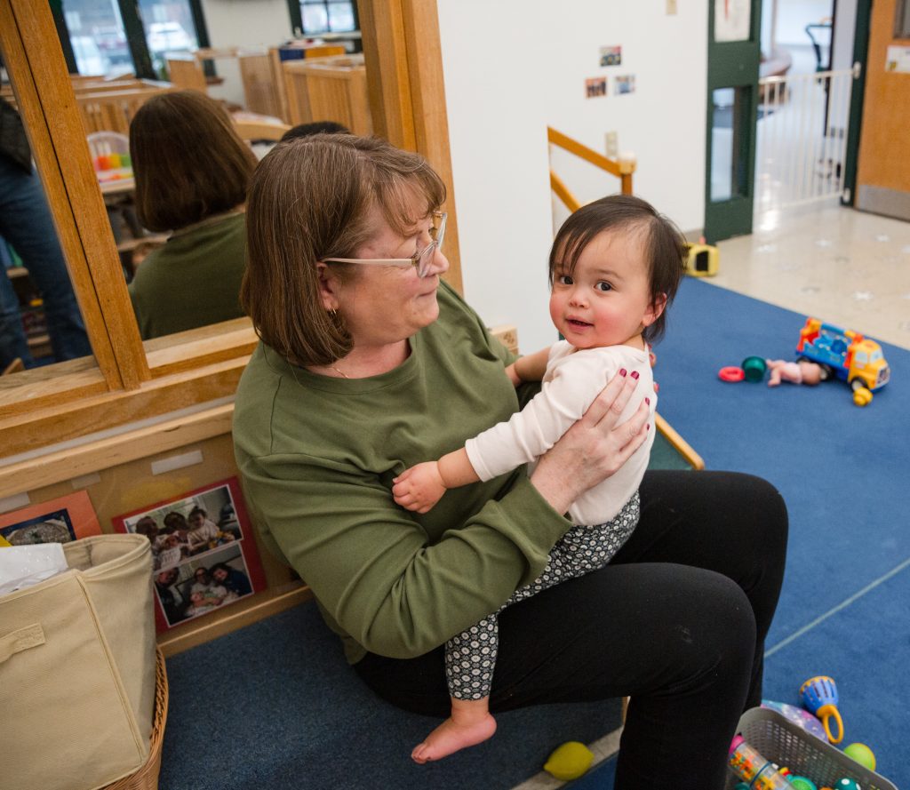 A woman wearing a green shirt and glasses sits close to the ground in a classroom, holding a baby on her lap.