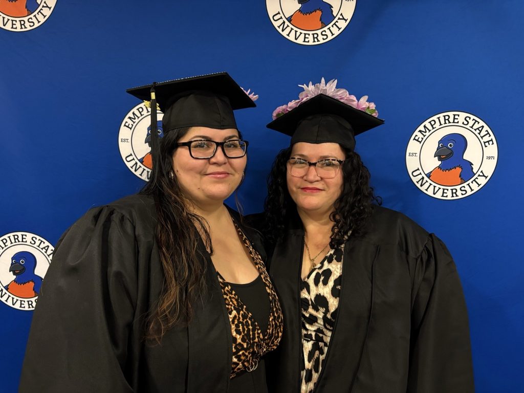 Two women wearing black glasses, black gowns and graduation caps, stand in front of a blue background bearing a logo that says Empire State University.